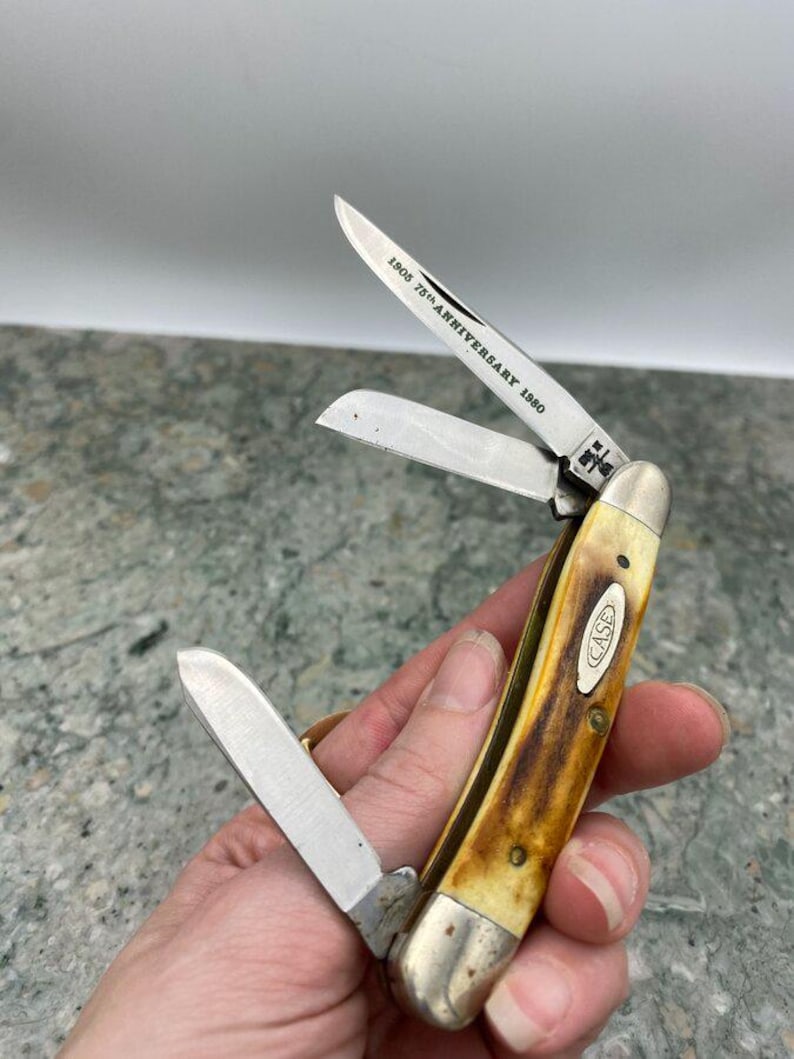 May include: A Case pocket knife with three open blades. The handle is a mix of yellow and brown, with a silver bolster and the Case logo. The blade reads "1905 75th Anniversary 1980".