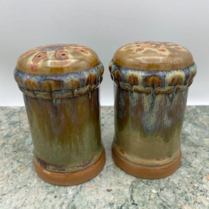 May include: A set of two ceramic salt and pepper shakers with a brown, green, and blue glaze. The shakers have a decorative design around the top and a textured base.