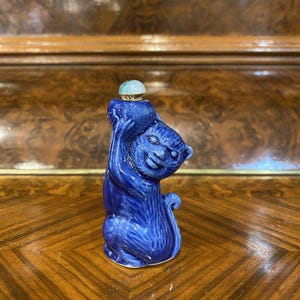 May include: A blue ceramic figurine of a cat with a light blue and white stone stopper on its head. The figurine is sitting on a wooden surface.