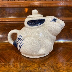 May include: A ceramic teapot shaped like a rabbit, with a cream-colored, crackled glaze. The rabbit teapot has blue accents on the ears, tail, and a floral design. The lid has a small knob.