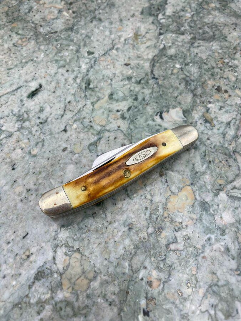 May include: A closed Case pocket knife with a brown and cream handle and silver-coloured bolsters. The knife has a single blade and the Case logo is visible. It rests on a speckled grey surface.