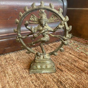 May include: A bronze statue of a dancing deity, framed by a circular halo with hand-like projections. The figure is in a dynamic pose, with multiple arms and intricate details. The statue is set on a square base and rests on a textured, brown surface.