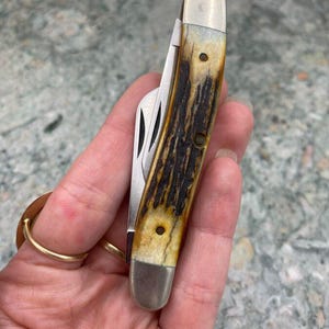 May include: A closed pocket knife with a handle displaying a brown and tan pattern, silver-coloured metal bolsters, and a stainless steel blade. The handle's textured design is visible.