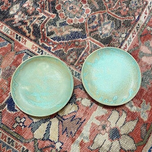 May include: Two teal blue ceramic bowls with a light brown glaze on a red, blue, and white patterned rug.
