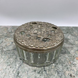 May include: A clear glass jar with a silver lid featuring an ornate floral design. The lid has small holes for ventilation.