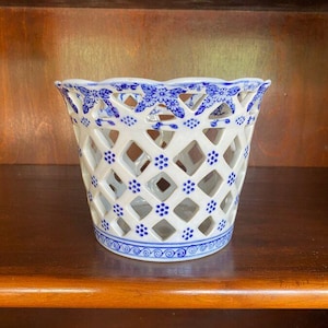 May include: White ceramic basket with a lattice design and blue floral accents. The basket has a scalloped rim and a decorative blue pattern around the base. The openwork design allows for air circulation and adds a touch of elegance.