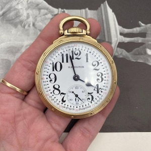 May include: A gold-toned Hamilton pocket watch with a white face and black numerals. The watch features a subdial and a loop for a chain. The watch hands point to approximately 11:00. The watch is held in a hand.