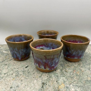 May include: Four small ceramic cups with a brown glaze and purple and blue accents. The cups are arranged in a square shape on a light gray surface.