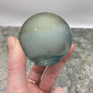 May include: A round, glass object with a gradient of colors, transitioning from a pale green at the top to a blue-green at the bottom. The glass has a slightly textured appearance and a small opening at the base. The object is held in a hand.