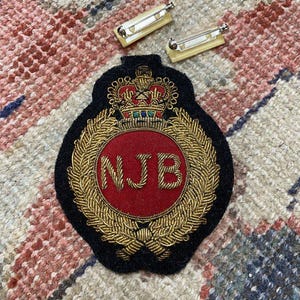 May include: A black and red embroidered patch with gold-colored detailing, featuring a crown and the letters "NJB". Two pin backs are visible above the patch. The patch is likely a decorative emblem or insignia.