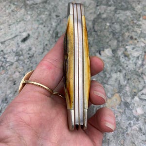 May include: A closed pocket knife with a bone handle and silver-coloured metal accents. The handle has a natural, mottled pattern of brown and cream colours. The knife is held in a hand, showing the side profile of the closed blades.