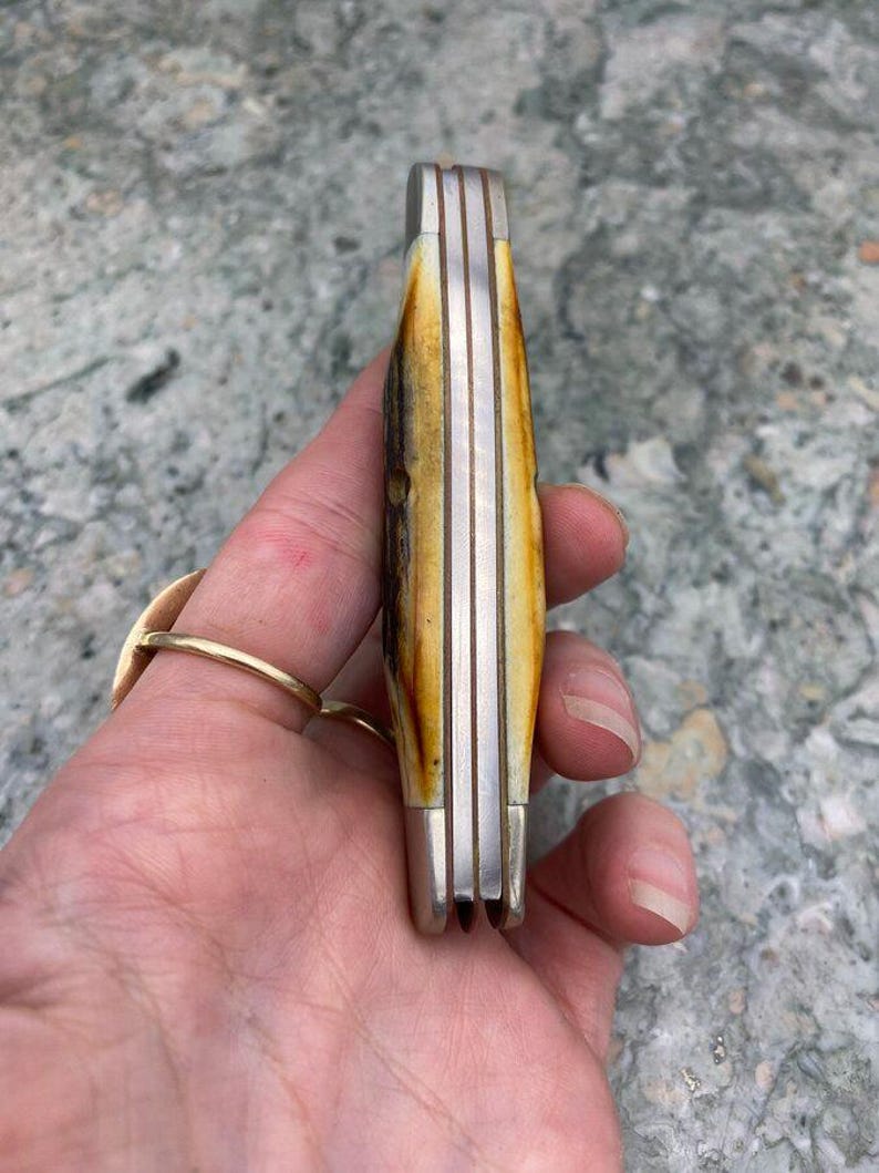 May include: A closed pocket knife with a bone handle and silver-coloured metal accents. The handle has a natural, mottled pattern of brown and cream colours. The knife is held in a hand, showing the side profile of the closed blades.