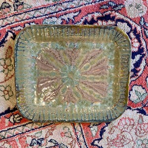 May include: Rectangular ceramic serving tray with a textured, glazed surface. The tray features a central design in shades of brown, green, and blue, with a radiating pattern. The edges have a raised, patterned border. A decorative piece for serving.