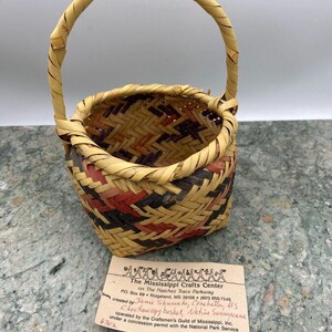 K&ouml;nnte beinhalten: Handgewebter Korb mit Henkel, mit einer Mischung aus nat&uuml;rlichen, roten und schwarzen Webmustern. Der Korb hat eine runde Form und einen stabilen Henkel. Ein Etikett lautet "Choctawegg basket, Native Swampcane".