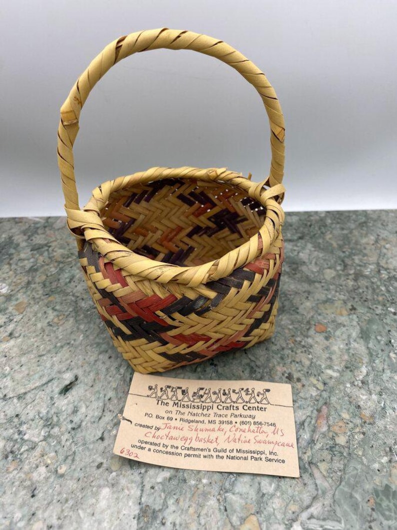 K&ouml;nnte beinhalten: Handgewebter Korb mit Henkel, mit einer Mischung aus nat&uuml;rlichen, roten und schwarzen Webmustern. Der Korb hat eine runde Form und einen stabilen Henkel. Ein Etikett lautet "Choctawegg basket, Native Swampcane".
