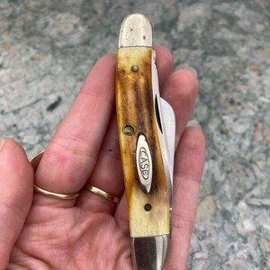 May include: A Case pocket knife with a brown and cream handle and a silver blade. The knife has a classic design with the "CASE" logo on the handle. The handle has brass rivets and a metal bolster.