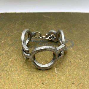 May include: A silver-toned metal bracelet with a series of connected circular and oval links. The bracelet has a vintage or antique appearance, with a slightly tarnished finish. The design features a chain clasp.
