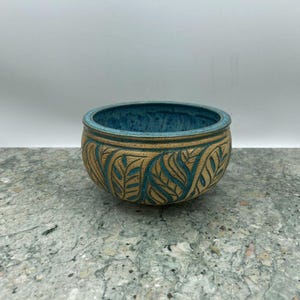 May include: A round ceramic bowl featuring a textured tan exterior with carved leaf patterns filled with a deep blue glaze. The interior of the bowl is finished in a matching deep blue glaze, creating a contrast between the earthy tan clay and the vibrant blue color.