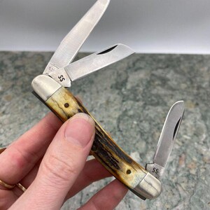 May include: A multi-blade pocket knife with three open stainless steel blades. The handle is a light brown with dark brown marbling. The knife has the initials "SS" on the metal. The blades are open, showing the sharp edges.