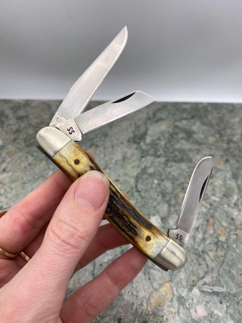 May include: A multi-blade pocket knife with three open stainless steel blades. The handle is a light brown with dark brown marbling. The knife has the initials "SS" on the metal. The blades are open, showing the sharp edges.
