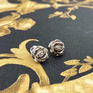 May include: A pair of silver-toned rose stud earrings. The earrings are detailed with delicate petals, and are set against a black background with a gold floral pattern. These earrings are a classic jewelry accessory.