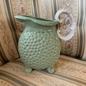 May include: A light green ceramic pitcher with a textured body and a white, ornate handle. The pitcher has a round base with three small feet. The handle is curved and detailed. The pitcher is likely for decorative use.