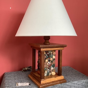 May include: A wooden table lamp with a white lampshade. The base of the lamp is made of wood and has a column of colorful stones in the center.