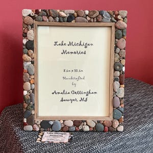 May include: A rectangular picture frame with a wooden border, embellished with various-sized stones. The frame holds a cream-colored paper with the text "Lake Michigan Memories" and "8 in x 10 in". Handcrafted by Amalia Cottingham, Sawyer, MI.
