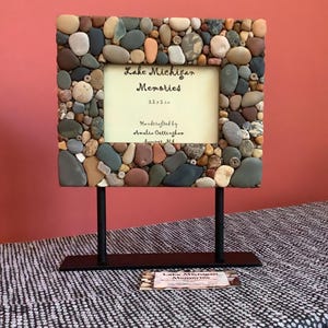 May include: A rectangular photo frame made of various sized, colorful stones. The frame holds a cream-colored insert with the text "Lake Michigan Memories" and the dimensions 3.5 x 5 in. It stands on a black metal stand.
