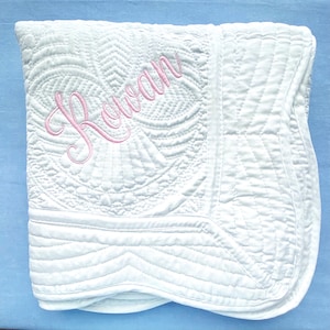 May include: White quilted baby blanket with a pink embroidered name "Rowan" in a cursive font.