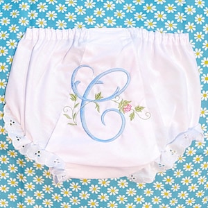May include: White baby bloomers with a light blue embroidered initial "C" and floral accents. The bloomers have a scalloped lace trim along the leg openings and a gathered waistband. The background is a blue fabric with white daisy patterns.