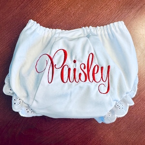 May include: Light blue baby bloomers with the name "Paisley" embroidered in red script. The bloomers have a ruffled, lace trim around the legs and waist, and are made of a soft fabric.