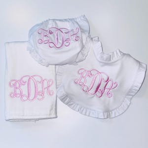 May include: A white fabric set consisting of a burp cloth, ruffled diaper cover, and a ruffled bib. Each item features a pink embroidered monogram with the letters BDA in a cursive script. The diaper cover has eyelet lace trim edges.