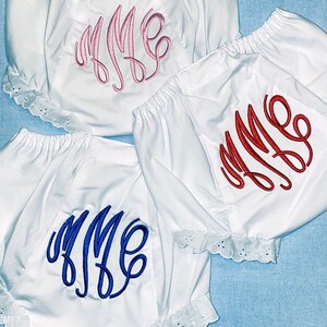 May include: White baby bloomers with embroidered monograms in pink, red, and blue. The bloomers have elastic waistbands and eyelet lace trim along the leg openings. The background is a light blue.