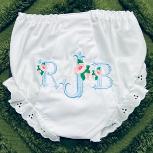 May include: White baby bloomers with an elastic waistband and a scalloped lace trim. The bloomers feature the embroidered initials "RJB" in light blue, each adorned with pink roses and green leaves.