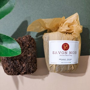 Peut inclure: Deux morceaux de savon noir africain. L'un est un bloc brun foncé à la texture rugueuse. L'autre est enveloppé dans du papier brun avec une étiquette indiquant "SAVON NOIR African Black Soap". L'étiquette indique également "100% naturel. Fait main".