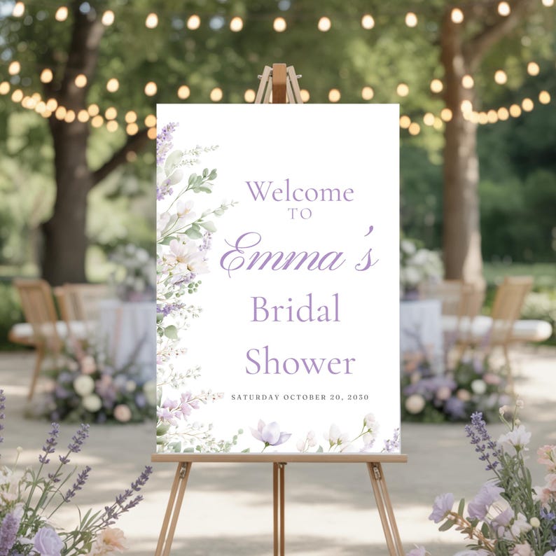 May include: A welcome sign for Emma's bridal shower. The sign is white with floral accents in shades of purple and green. The text reads 'Welcome to Emma's Bridal Shower' with the date 'Saturday October 20, 2030' at the bottom. The sign is displayed on a wooden easel outdoors.