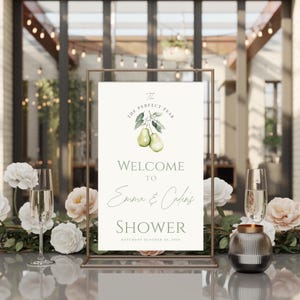 May include: A framed sign with the text "The Perfect Pear" and "Welcome to Emma & Cadie's Shower, Saturday October 20, 2030." The sign features pear illustrations and is set on a table with floral arrangements and champagne glasses.