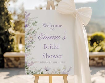 May include: A wooden easel holds a sign that reads "Welcome to Emma's Bridal Shower" in purple script. The sign is decorated with floral accents and a large white bow. The date "Saturday October 20, 2050" is printed at the bottom.