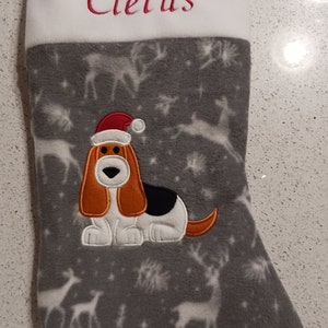 Puede incluir: Una media de Navidad gris y blanca con un puño blanco. El puño tiene el nombre "Cletus" bordado en rojo. La media tiene un fondo gris con renos blancos y copos de nieve. Un basset hound marrón y blanco con un gorro de Papá Noel rojo está bordado en la media.