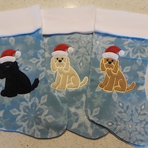 Può includere: Tre calze di Natale blu con polsini bianchi e motivi a fiocco di neve. Ogni calza presenta un cane di colore diverso con un cappello di Babbo Natale: nero, marrone chiaro e marrone.