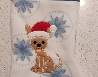 May include: A white fleece Christmas stocking featuring a blue cuff embroidered with the name Pixie. The body of the stocking has a light blue snowflake pattern and an embroidered tan dog wearing a red and white Santa hat in the center.