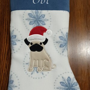 May include: A white Christmas stocking with a blue cuff and snowflake pattern. The stocking has a personalized name "Obi" embroidered on the cuff and a felt pug wearing a Santa hat.