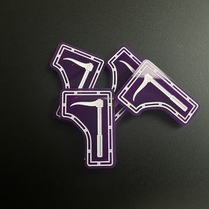 May include: A collection of purple and white scythe-shaped stickers. Each sticker features a white scythe design within a white-bordered, purple background. The stickers are scattered on a black surface.