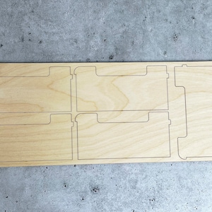 May include: A wooden organizer with five slots for holding papers or other items. The organizer is made of light-colored wood and has a simple, modern design.