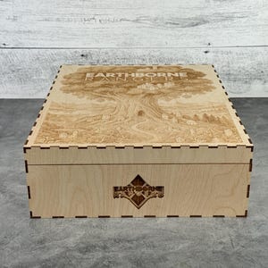 May include: A wooden box with a detailed engraved design on the lid, featuring a large tree and the text "EARTHBORNE RANGERS". The box has a decorative diamond-shaped emblem on the front and is made of light-colored wood.