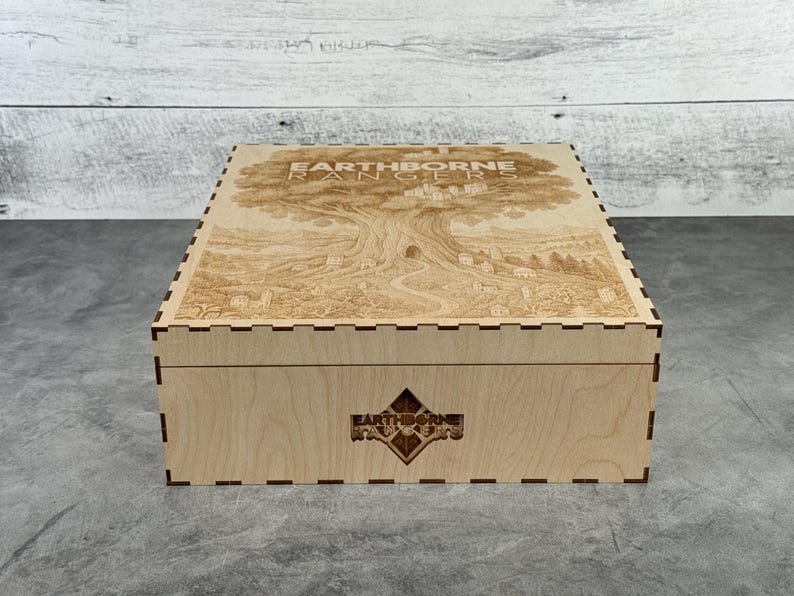 May include: A wooden box with a detailed engraved design on the lid, featuring a large tree and the text "EARTHBORNE RANGERS". The box has a decorative diamond-shaped emblem on the front and is made of light-colored wood.