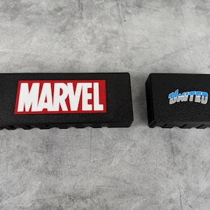 May include: Two black rectangular boxes with a textured surface. The box on the left has the word "MARVEL" in white letters on a red background. The box on the right has the word "UNITED" in blue letters on a white background.