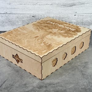 May include: A light-colored wooden box with a detailed engraved design on the lid, featuring a tree and the text "EARTHBORNE RANGERS". The box has decorative cutouts on the sides and a geometric pattern.