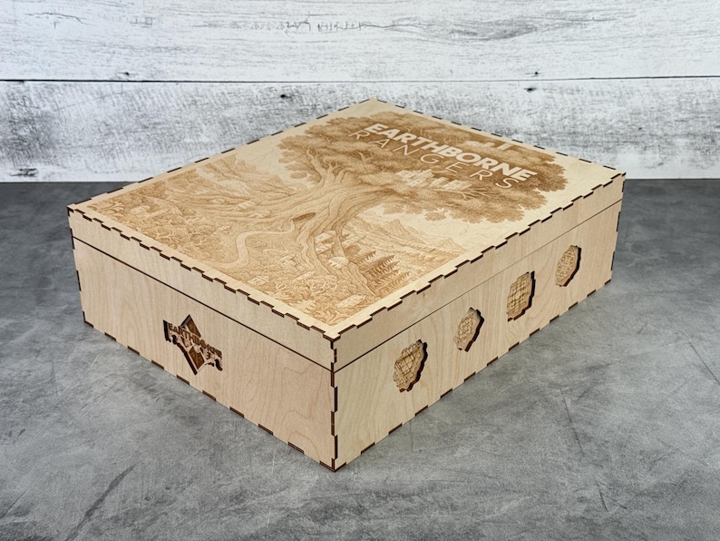 May include: A light-colored wooden box with a detailed engraved design on the lid, featuring a tree and the text "EARTHBORNE RANGERS". The box has decorative cutouts on the sides and a geometric pattern.